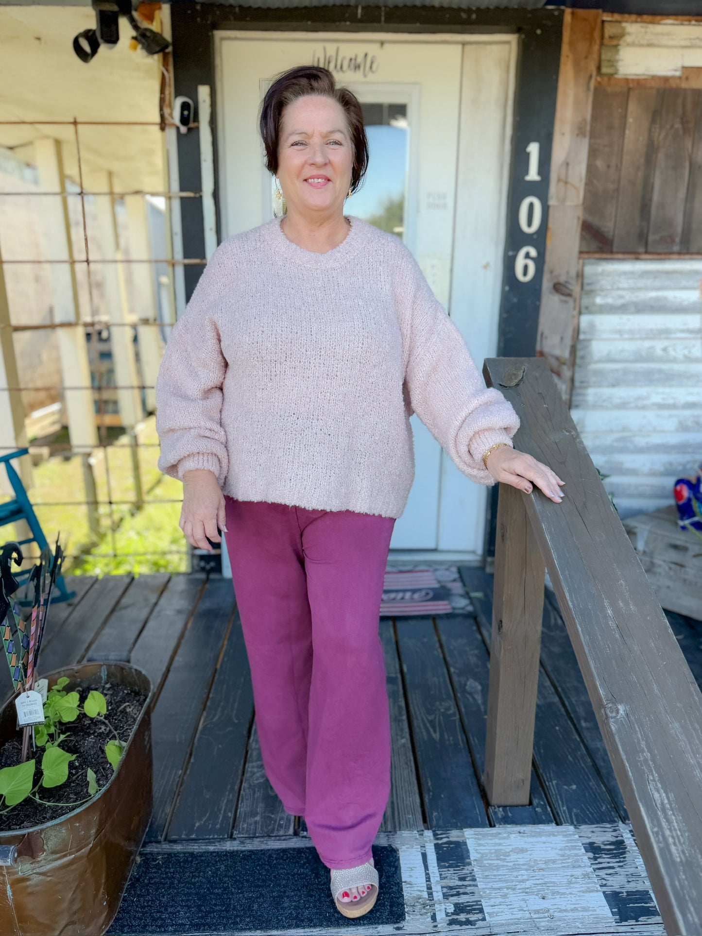 Woman standing on a wooden porch wearing a pink sweater and purple pants.