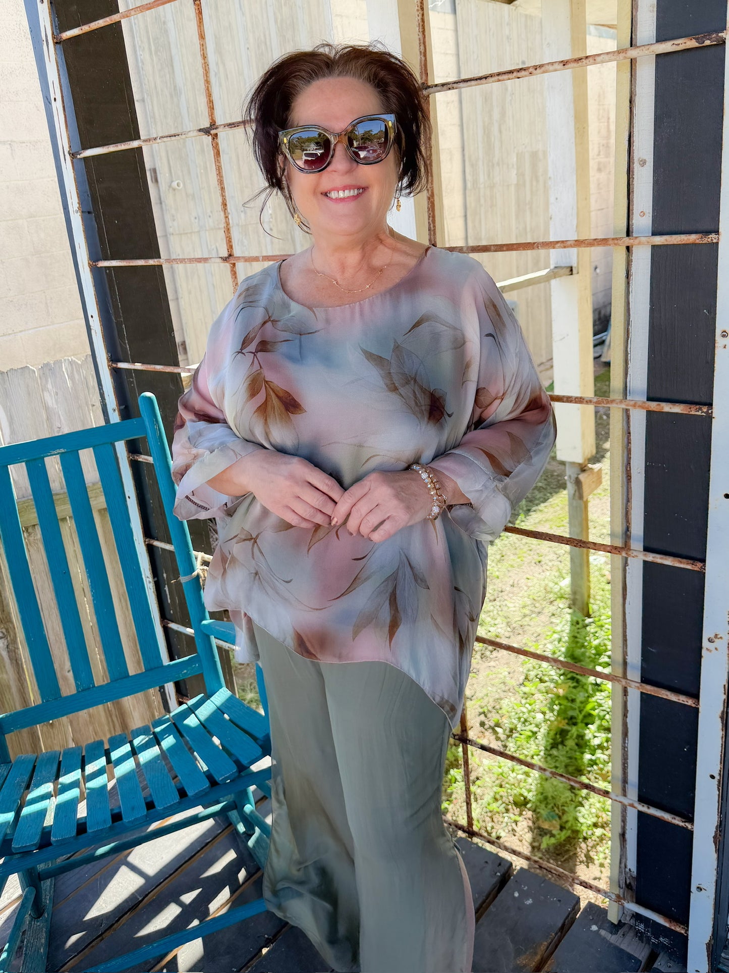 Woman wearing a patterned top and sunglasses standing on a wooden deck.