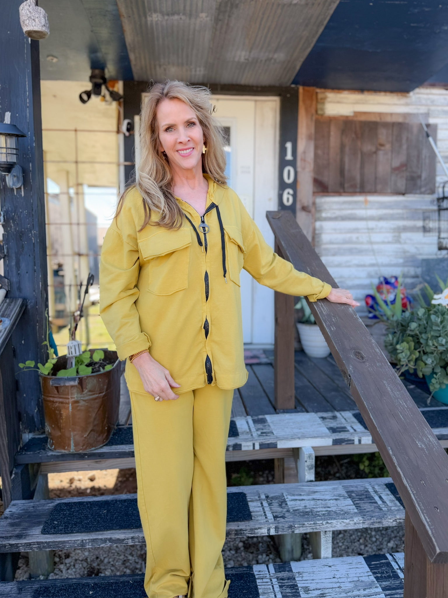 Woman in a yellow raincoat standing on a porch with plants and a house number visible.