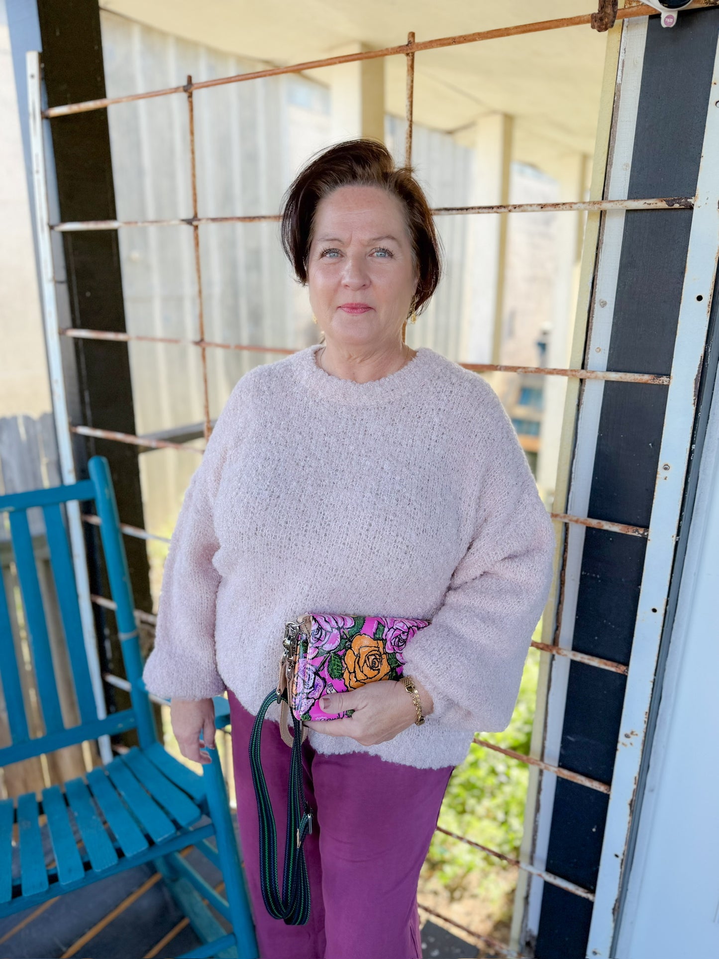 Woman in a pink sweater and purple pants holding a colorful clutch on a porch.