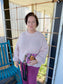 Woman in a pink sweater and purple pants holding a colorful clutch on a porch.