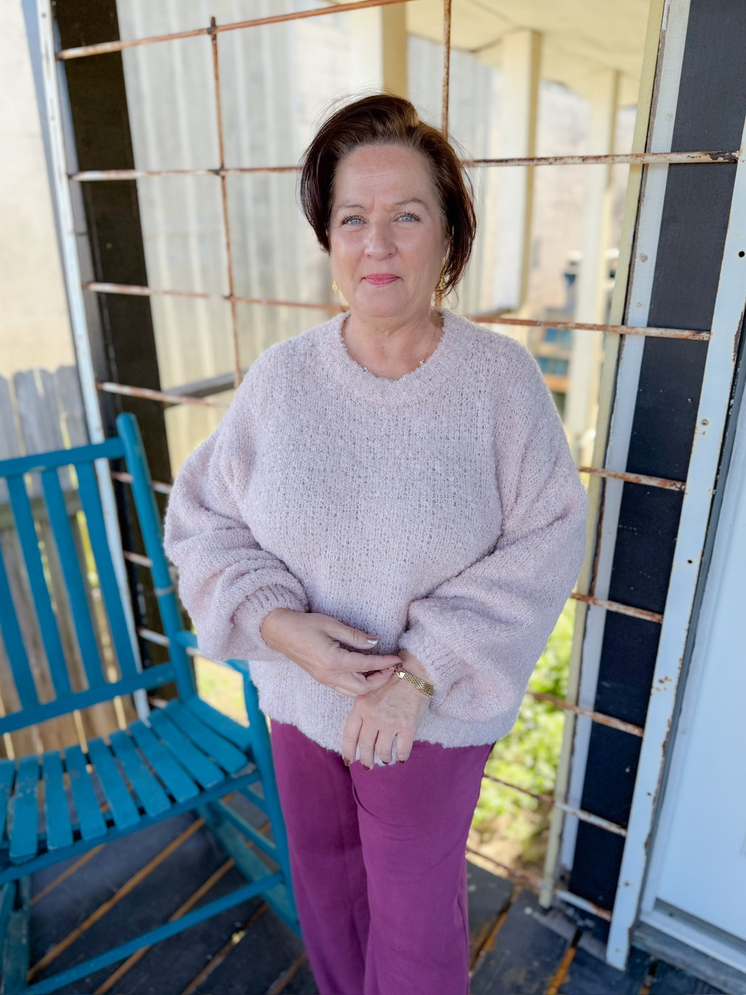Woman standing on a porch wearing a light pink sweater and purple pants.