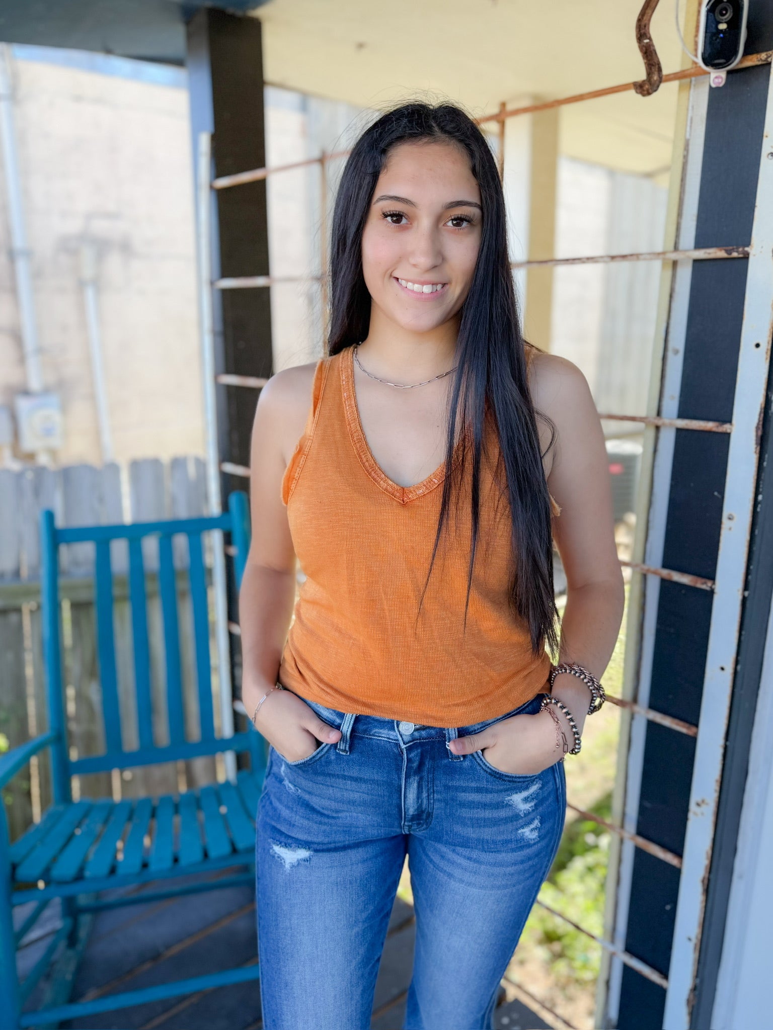 Woman wearing an orange tank top and blue jeans standing on a porch.