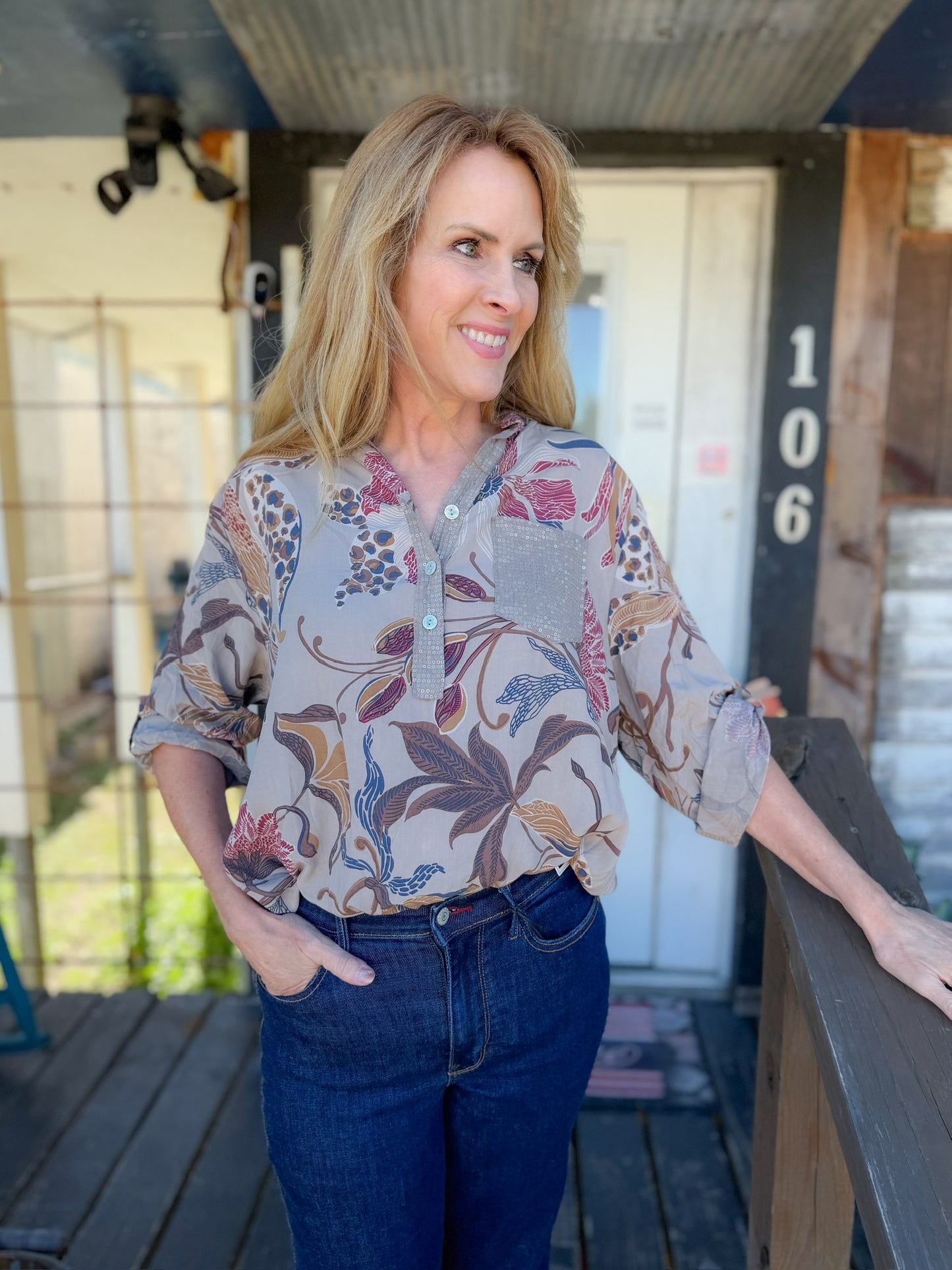 Woman standing on a wooden deck with a floral shirt and jeans, with a building number '106' in the background.